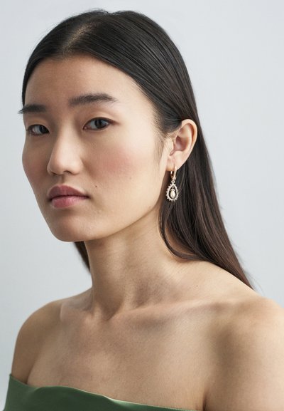 Goldene Tropfohrringe mit einem Perlenmittelstück, die über feine Details und eine Tropfenform verfügen, getragen mit einem schulterfreien grünen Oberteil.