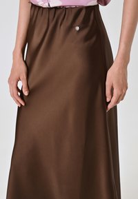 Bruine satijnen rok met elastische tailleband en een klein zilveren detail aan de voorkant, met een gladde, strakke textuur.