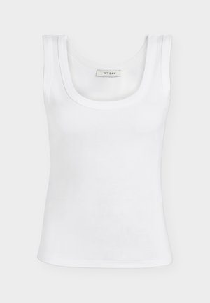 Kvinde med kort krøllet hår iført hvid tanktop, denimshorts, sort-hvide sneakers, bærende stor sort skuldertaske.