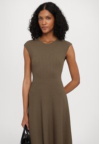 Ribbed, olive green dress with a round neckline and cap sleeves. Flared skirt design with a smooth texture. Held with a black handbag.