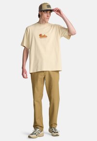 Ελαφρύ μπεζ βαμβακερό t-shirt με πορτοκαλί γραφικό, συνδυασμένο με καφέ παντελόνι. Το μοντέλο φοράει καπέλο σε καφέ απόχρωση και sneakers με σχέδια.