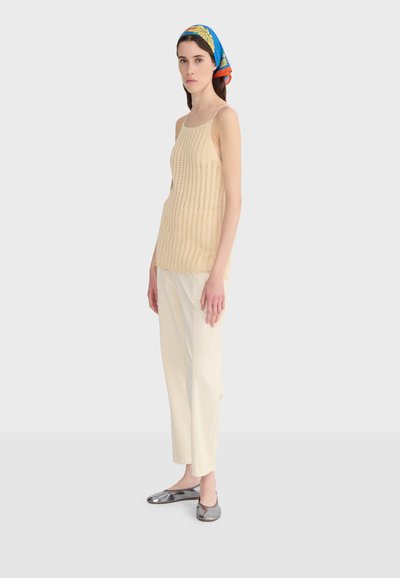 Femme portant un haut en maille beige sans manches, un pantalon crème, des ballerines métalliques argentées, et un foulard coloré avec des motifs bleus, jaunes et orange, debout de profil.