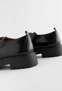 Zapatos de piel negra con un acabado brillante, que cuentan con una suela gruesa y texturizada y detalles decorativos de brogue a lo largo del borde superior.