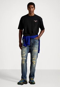 Μαύρο βαμβακερό μπλουζάκι, μπλε πουλόβερ δεμένο στη μέση, φθαρμένο μπλε τζιν με ανοιχτές κηλίδες και πολύχρωμα αθλητικά παπούτσια.