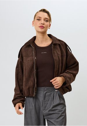 Ruda suede bomber tipo striukė su užtrauktuku, laisvas fasonas ir elastingomis rankovėmis. Nešiojama ant tamsiai rudos palaidinės, derinama su pilkai dryžuotais kelniais.