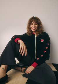 Junge Frau mit lockigem Haar sitzt lächelnd, trägt eine schwarze Jacke mit roten Herz-Patches und schwarze Hosen mit weitem Bein auf beigem und schwarzem Boden.