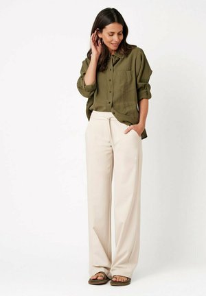 Woman standing wearing an olive green button-up shirt with rolled sleeves, beige wide-leg pants, and brown sandals.