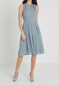 En ljusblå ärmlös klänning med en texturerad överdel, dekorerad med paljetter, och en flödande plisserad kjol. Silverfärgade klackar fulländar looken.