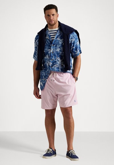Cămașă cu model floral, de culoare albastră, cu mâneci scurte, pulover bleumarin aruncat pe umeri, șorturi de baie roz deschis și încălțăminte casual de culoare bleu închis.