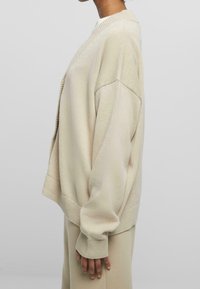 Beige oversized stickad tröja med V-ringning, sänkta axlar, ribbade manschetter och strukturerat mönster, matchad med beige byxor.