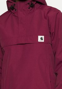 Veste à capuche couleur bordeaux avec une poche avant, des cordons de serrage ajustables et un patch logo blanc. Fabriquée en matière lisse et légère.