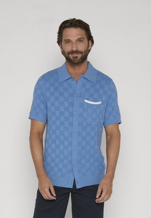 Man met baard die een blauw geruit overhemd met korte mouwen draagt, met een wit detail op het borstzakje, poseert tegen een grijze achtergrond.