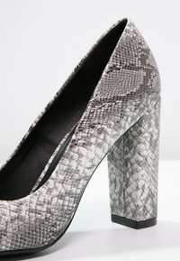 Scarpa con tacco alto in stampa serpente, caratterizzata da un tacco a blocco spesso, punta affusolata e materiale testurizzato con motivo grigio e nero.