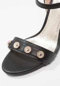 Sandale en cuir noir avec une bride comportant quatre clous décoratifs ronds dorés et une semelle intérieure métallique texturée.