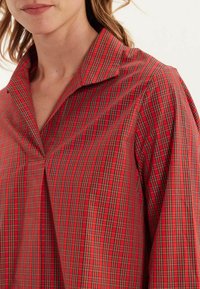 Chemise à carreaux rouges avec col pointu et coupe ample. Présente des lignes verticales et horizontales, un tissu texturé et un détail de pli à l'avant.