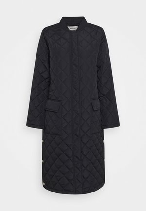 Long manteau noir matelassé avec col montant, présentant une couture diagonale, deux poches avant et des accents à pression en bas.