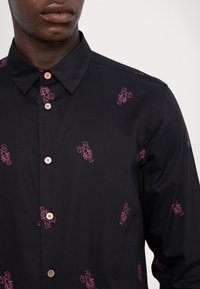 Homme portant une chemise noire à boutons avec des motifs de homards roses, montrant le col et le haut du torse sur un fond uni.