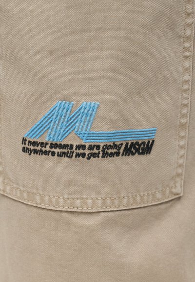 Tissu beige avec broderie bleue "ML" et texte noir sur la poche indiquant "On n'a jamais l'impression d'aller quelque part avant d'y arriver MSGM".