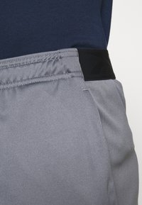 Grå träningsshorts med elastisk midja, kontrasterande svarta detaljer, slät textur och sidofickor, uppställda mot blått tyg.