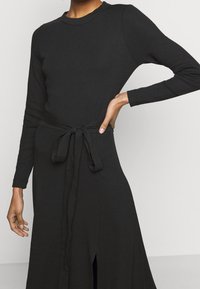 Vestito lungo nero a maniche lunghe con corpino aderente, fascia in vita legata e gonna fluida. Tessuto morbido con una sottile texture e dettaglio con spacco laterale.