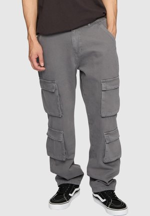 Person iført grå cargobukser med flere klaplommer og sorte sneakers med hvide såler, stående mod en ensfarvet baggrund.