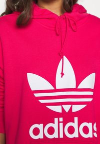Osoba v jasně růžové mikině Adidas s bílým trojlístkovým logem a šňůrkami uvázanými do mašle na krku.