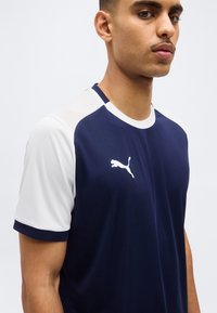 Jeune homme portant un maillot de sport Puma bleu marine et blanc, visible de la poitrine vers le haut, sur un fond uni clair.
