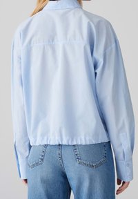 Lyseblå, oversized skjorte med en glat tekstur og lange ærmer. Har en spids krave og manchetter med knapper samt en rynket kant.