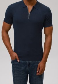 Marinblå ribbad kortärmad polotröja med dragkedja vid kragen; slät textur och figurnära design, kombinerad med mörkblå denimjeans.