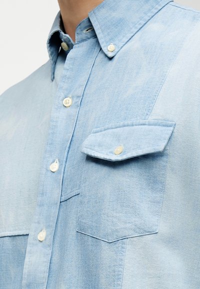 Γαλάζιο τζιν πουκάμισο με τσεπάκι στο στήθος που κλείνει με κουμπί, γιακά με κουμπιά και λευκά κουμπιά κατά μήκος του μπροστινού μέρους.
