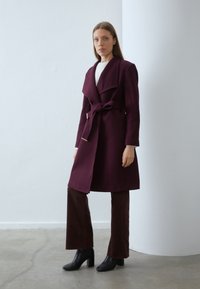Cappotto di lana bordeaux con ampi rever e una cintura annodata in vita, abbinato a pantaloni in corduroy marrone e stivaletti neri con tacco, su uno sfondo neutro.