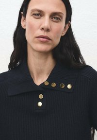 Donna con capelli lunghi e scuri che indossa un maglione a coste nero con colletto e sei bottoni rotondi dorati vicino al colletto.