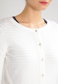 Valkoinen neuletakki, joka on valmistettu pehmeästä neuloskankaasta. Siinä on teksturoidut nyppykoristelut, pyöreä pääntie ja napitus läpinäkyvin napin.