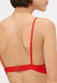 Červená podprsenka s tenkými nastaviteľnými ramienkami a háčkovým zapínaním. Hladká tkanina s minimalistickým dizajnom. Pohľad zozadu zdôrazňuje detaily pásu a ramienok.