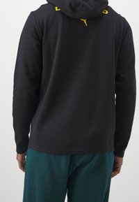 Persona che indossa un hoodie nero a maniche lunghe con un logo Puma giallo sul collo e pantaloni turchesi, visibile da dietro.