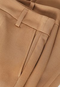 Kalhoty z hladkého béžového materiálu, s předními záhyby a plochým pasem. Stehy jsou viditelné podél okrajů.