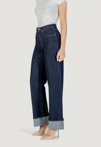 Jeans de denim azul de talle alto y pernera ancha con costuras rojas y dobladillos enrollados. Combinados con zapatos de tacón beige y una blusa blanca ajustada.