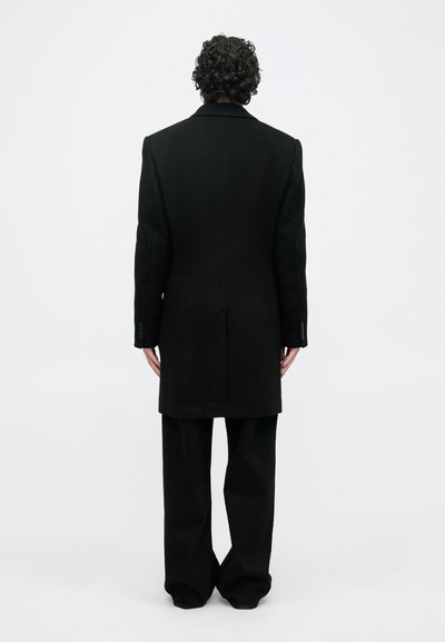 Άτομο με σγουρά μαλλιά στέκεται με την πλάτη γυρισμένη, φορώντας ένα μαύρο μακρύ παλτό και φαρδιά μαύρα παντελόνια, μπροστά από ένα λευκό φόντο.