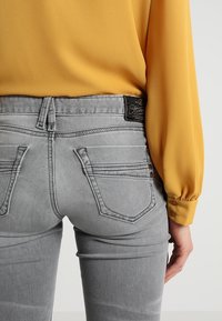 Šedé džíny z denimu s přiléhavým střihem, pěti kapsami a šitými detaily, spárované s žlutou košilí s dlouhým rukávem.