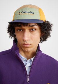 A sapka színes dizájnnal rendelkezik, zöld, narancssárga és bézs panelekből áll. Az elülső részen kék hímzéssel szerepel a "Columbia Sportswear Company" felirat.