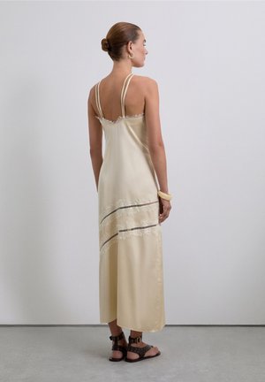 Vestido de satén tipo slip en amarillo pálido con tirantes finos. Presenta detalles de flecos en capas y rayas horizontales cerca del dobladillo. Llevado con sandalias marrones.