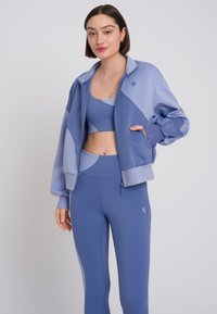 Set atletico leggero in diverse sfumature di blu. Include una giacca con zip e leggings aderenti con una finitura liscia e un branding minimal.