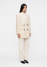 Διπλόμπελο ελαφρύ μπεζ σακάκι με μαύρα κουμπιά, συνδυασμένο με μπεζ παντελόνι φαρδιάς γραμμής και μαύρες μυτερές γόβες, με δομημένους ώμους.