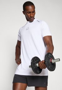 Weißes Sport-T-Shirt mit kurzen Ärmeln und grauem Logo, kombiniert mit schwarzen Shorts. Die Person hält eine schwarze Hantel mit silbernen Akzenten.