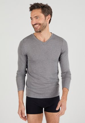 Glimlachende man met kort bruin haar en baard, die een grijs langarmig shirt met V-hals en zwarte boxers draagt tegen een effen achtergrond.