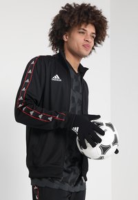 Zwarte sportjack met rode strepen, contrasterend adidas-logo, zwarte handschoenen en een voetbal. Model draagt een donker gestreept shirt eronder.