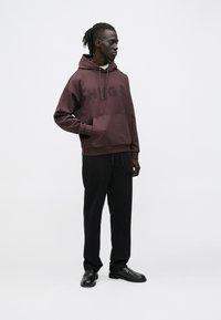 Uomo con dreadlocks indossa una felpa con cappuccio color bordeaux con la scritta "HUGO", pantaloni neri e scarpe nere, in piedi davanti a uno sfondo chiaro uniforme.