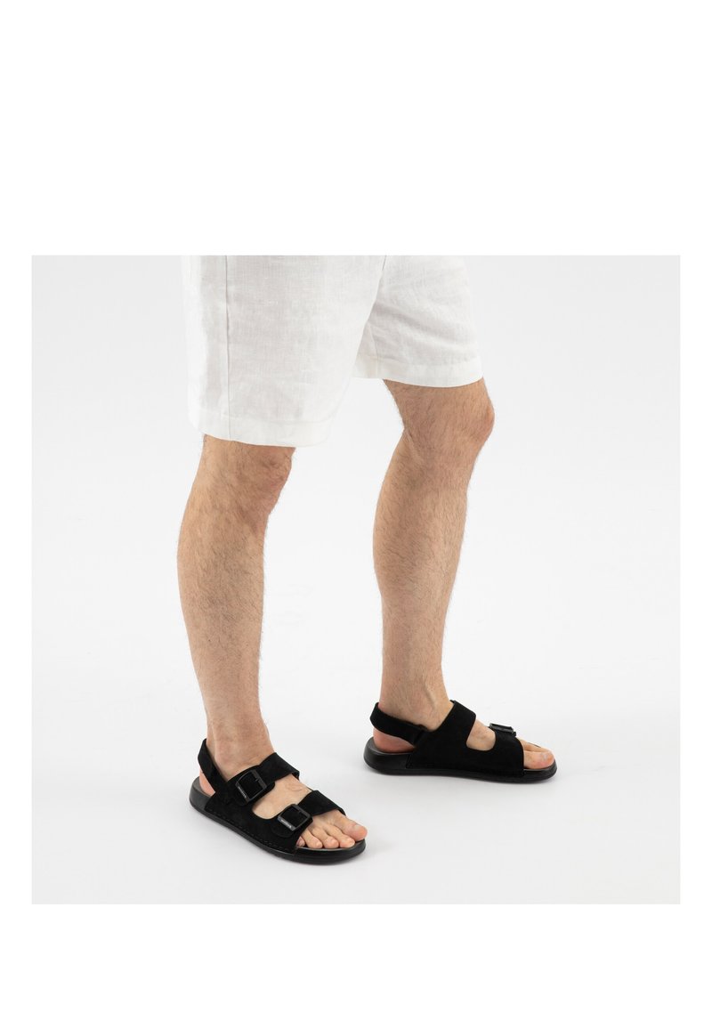 Beine in weißen Shorts und schwarzen Sandalen mit offenen Zehen und doppelten Schnallenriemen, die auf einer schlichten weißen Fläche stehen.