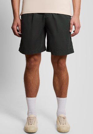 Homme portant un short noir longueur genoux, des chaussettes blanches et des baskets en toile beige, debout devant un fond uni.