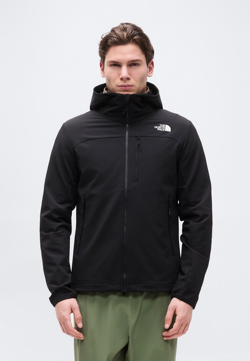 Jeune homme portant une veste à capuche noire zippée avec le logo "The North Face", debout devant un fond clair uni.